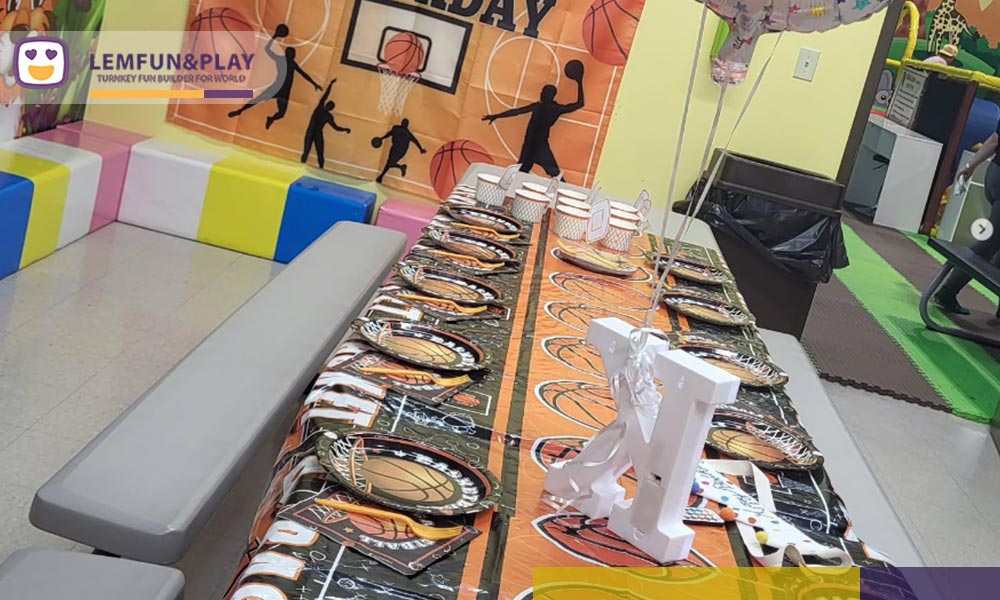 A photograph showing a decorated party area with several small tables and chairs arranged with colorful tablecloths, party hats, and balloons, in front of a large, colorful mural of jungle animals and a 'HAPPY BIRTHDAY' banner.