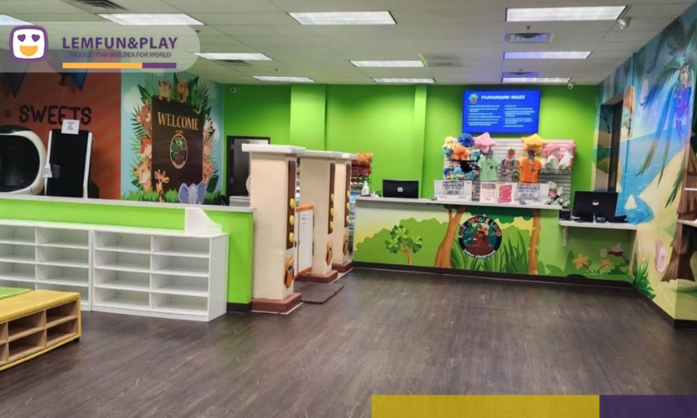 A photograph of a clean, modern reception desk and check-in counter at Lemfun Play Land, featuring a white curved front, a stylized tree logo on the wall behind, and point-of-sale systems, showing the facility entrance.