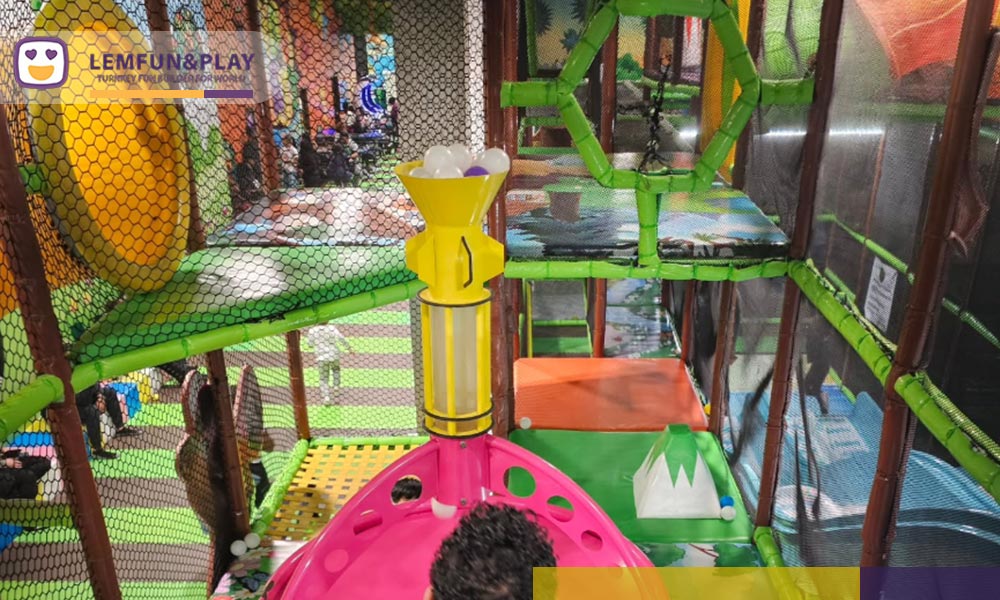 A detailed photograph of an interactive ball machine, a transparent pneumatic tube system where children use air pressure to launch and direct plastic balls through a maze of pipes on a wall.